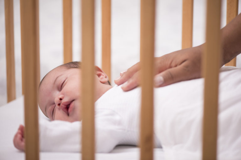 A baby asleep in their cot with a comforting hand on their chest.