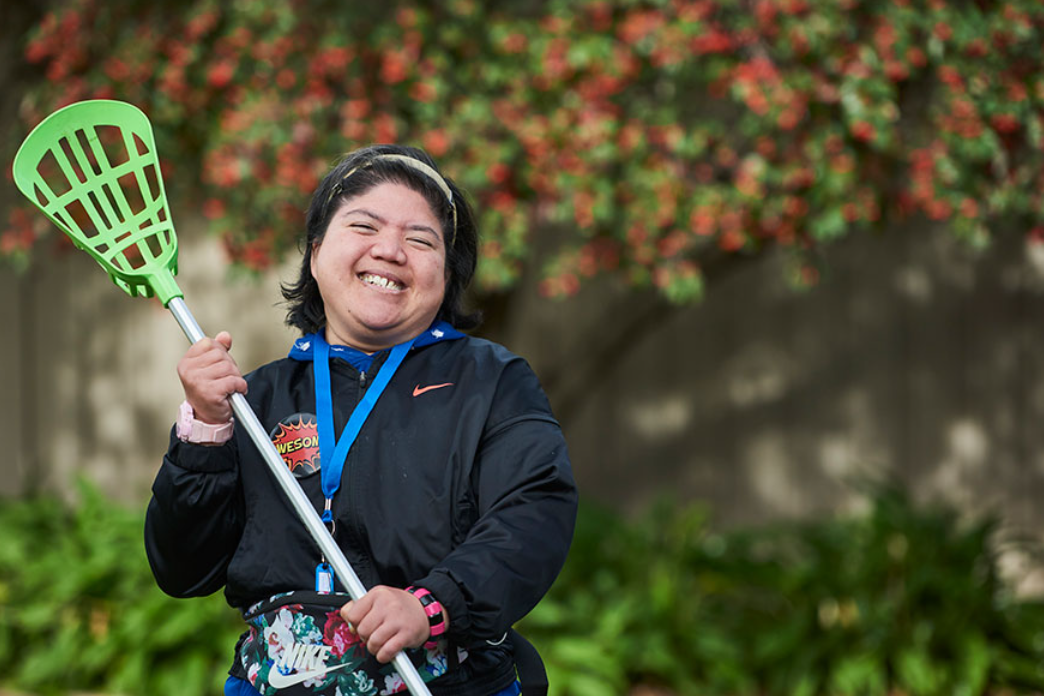 A young female smiling while holding a lacrosse bat.
